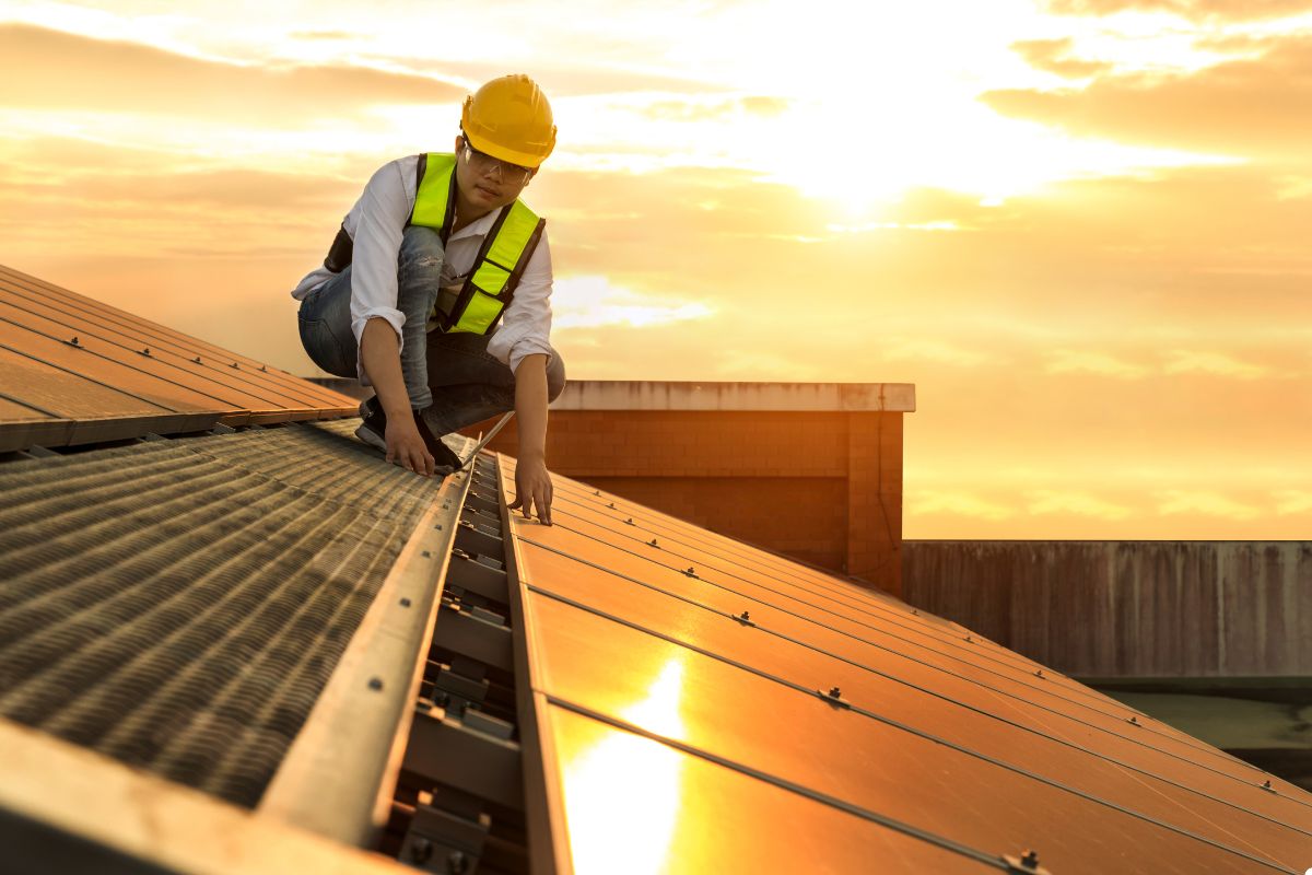 A worker in a hard hat and safety vest is engaged in the installation of a solar panel under clear blue skies.