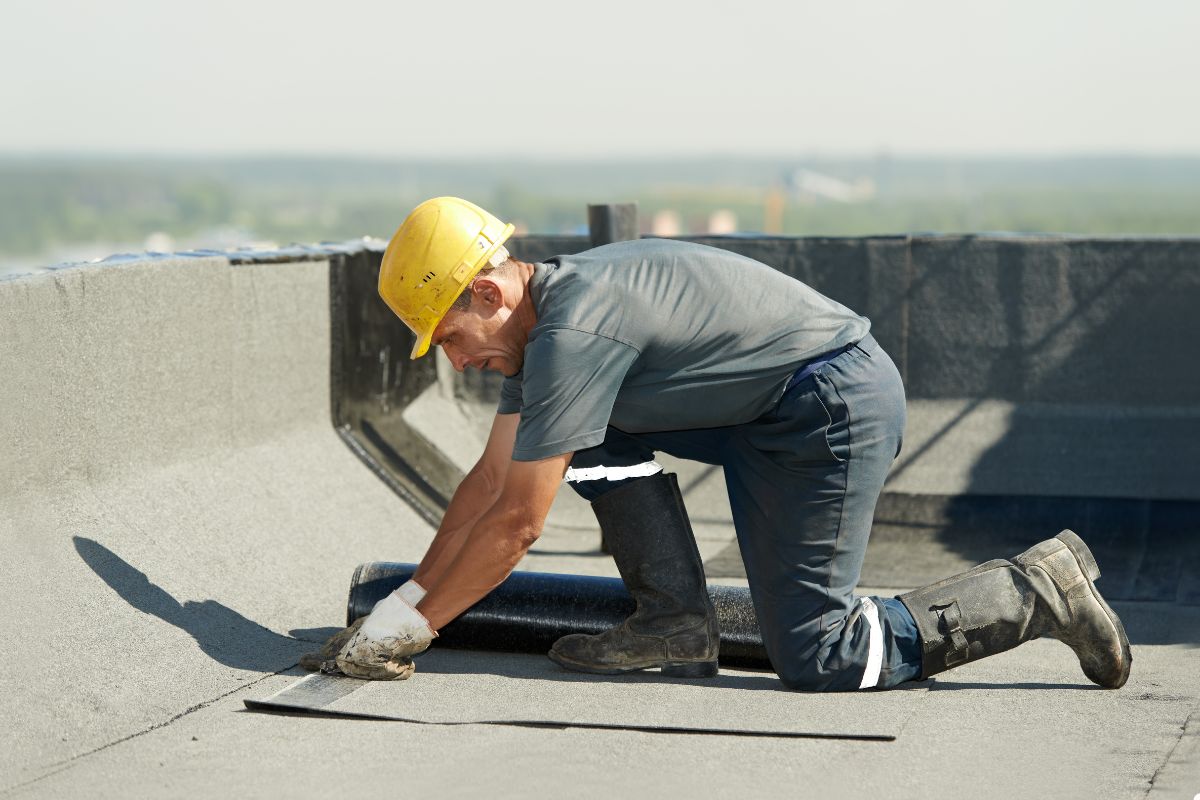 A man wearing a hard hat and yellow safety vest is lying on a roof, inspecting or working on the structure.