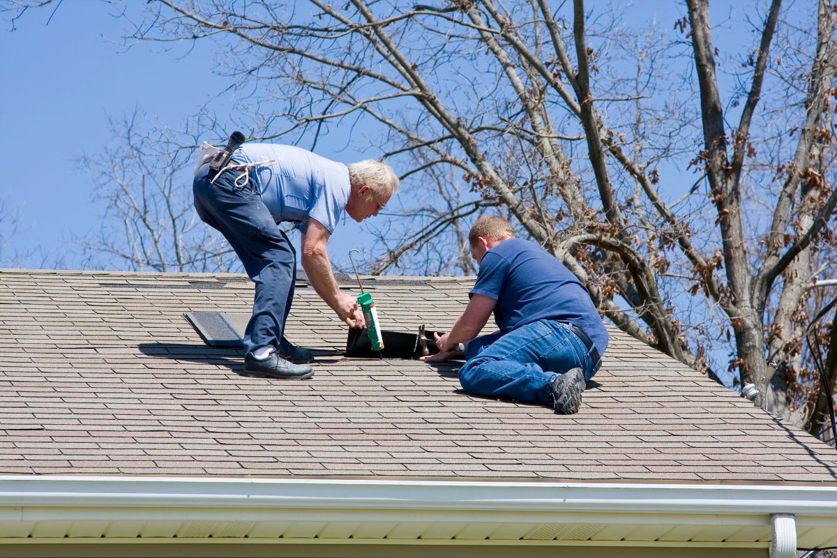 Two men work on a sloped roof, applying sealant around a vent, with bare trees and a clear blue sky in the background.