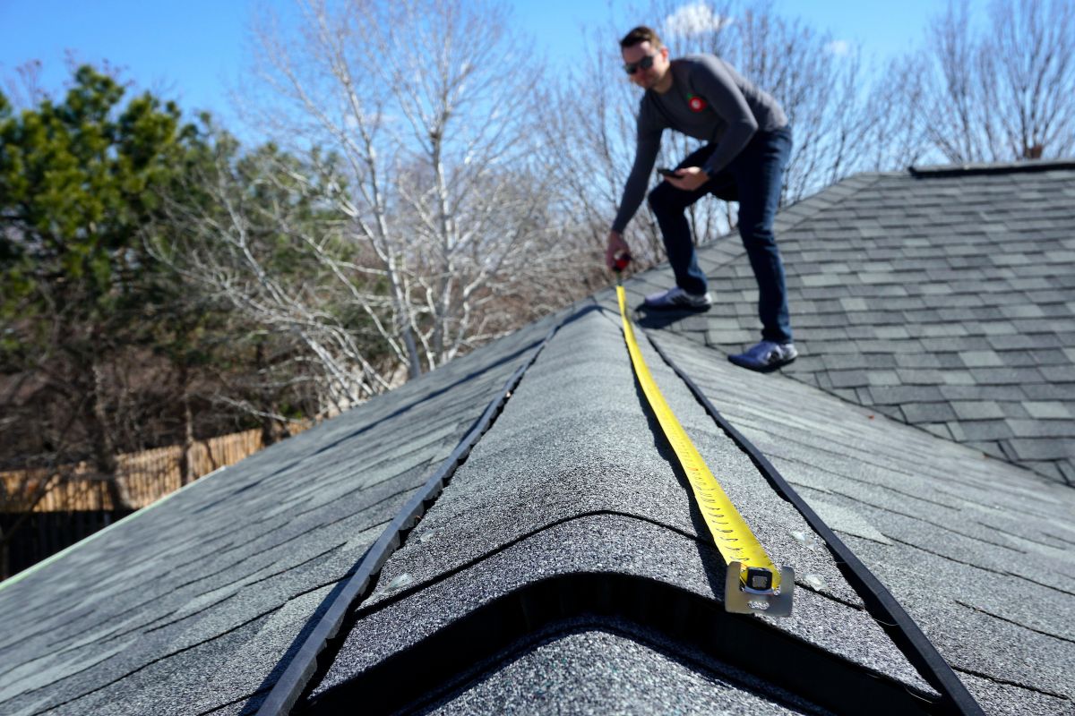 A person measures the roof's peak with a yellow tape measure while standing on a sloped shingle roof under a clear blue sky.